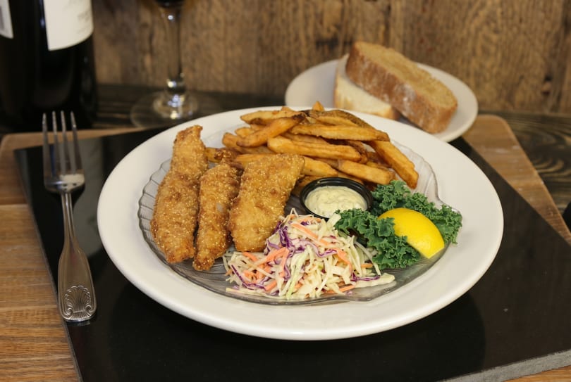 A plate of golden-brown fish and chips with coleslaw, tartar sauce, and a lemon wedge.