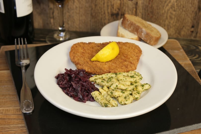 A schnitzel with a lemon wedge, red cabbage, and spaetzle on a white plate.