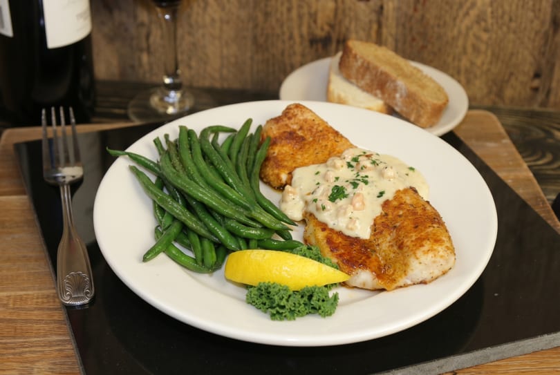 Pan-seared fish with creamy sauce, green beans, lemon, and bread on a rustic table.