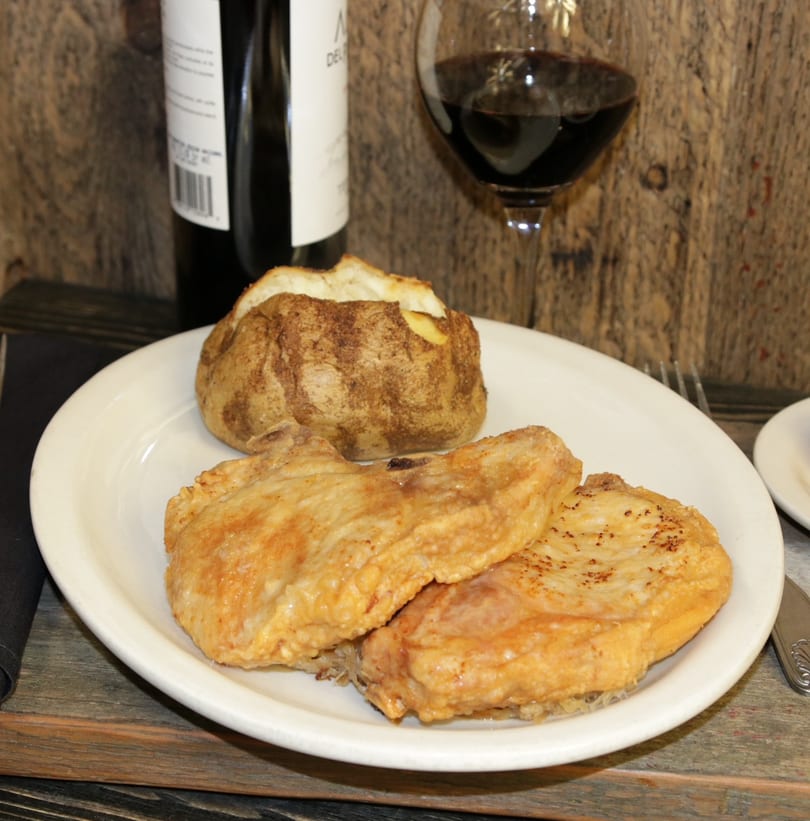 Two golden-brown pork chops and a baked potato on a white plate, with red wine in the background.