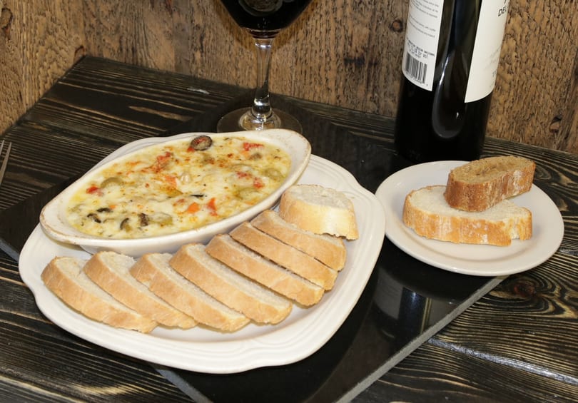 Baked dip with vegetables and cheese, served with sliced baguette and a glass of red wine.