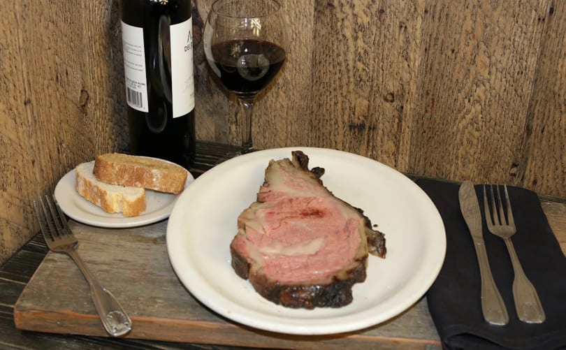 A slice of prime rib, bread, and red wine on a rustic wooden table.