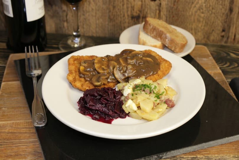 A plate of schnitzel with mushroom gravy, red cabbage, and potato salad, with wine and bread.